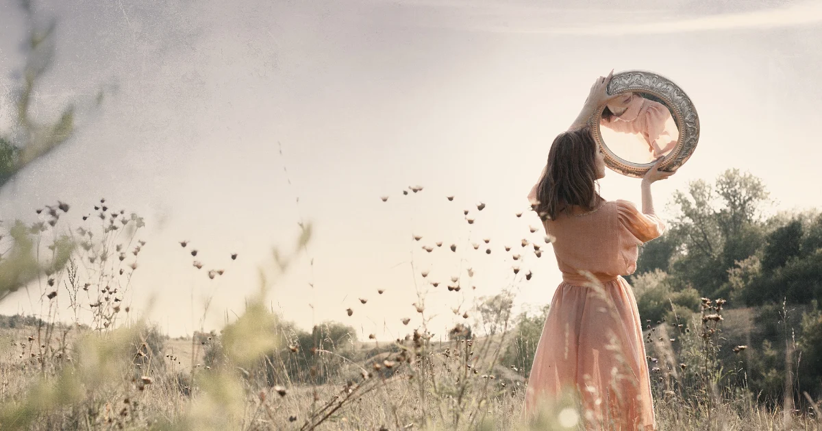 A girl standing in the middle of field looking in a mirror
